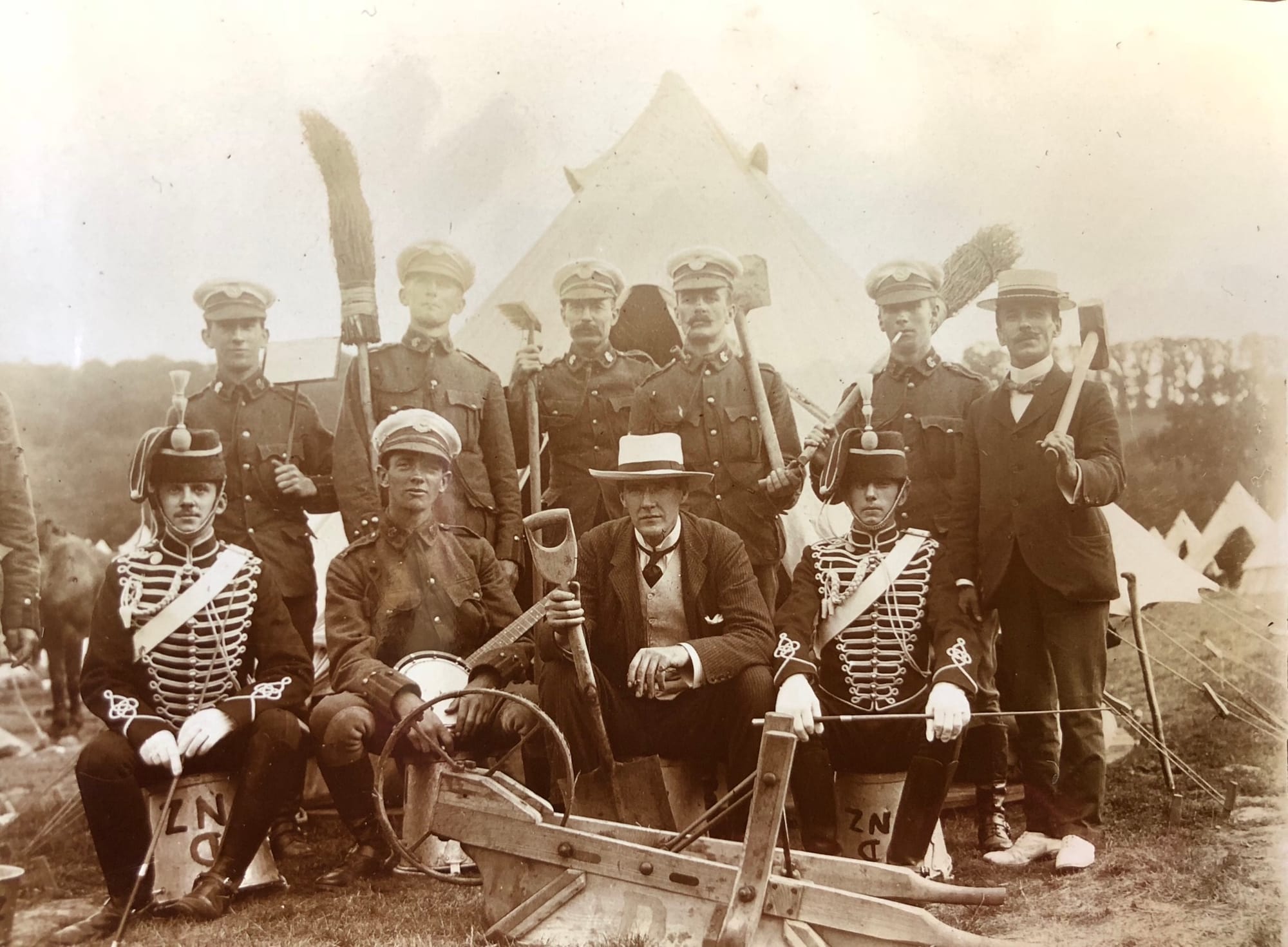 Fatigue party of New Zealanders of ‘D’ Squadron (British African) King’s Colonial’s with visitors from ‘D’ (Chesham) Squadron, The Buckinghamshire Imperial Yeomanry (Royal Buckinghamshire Hussars) and civilians in 1903. The 'D NZ' painted on the upturned pales designate these New Zealand Troopers as belonging to ‘D’ Squadron.  ‘D’ Squadron was the British African Squadron but the 3rd Troop was comprised of the New Zealanders who had originally been part of ‘C’ Squadron (Australasian) in 1902. The Troopers (front left and front third from left) are still wearing the ‘C’ Squadron (Australasian) headdress badge (and an NCO’s arm badge, Figure 151) prior to the fern badge of the New Zealand Squadron being introduced in 1904. Other Troopers (rear left-hand two and front right) are wearing the ‘D’ Squadron (British African) headdress badge. There is a very good description of the Full Dress uniform of the Royal Bucks Hussars (not Imperial) in 'The Yeomanry Force at the 1911 Coronation' by R. J. Smith and R. G. Harris (Picton Publishing, Chippenham, 1988) which matches the uniform worn by the two Buckinghamshire Imperial Yeomanry. A regulation or low pattern Hussars busby with scarlet bag, white lines and white plume. Jacket invisible green with scarlet collar and white chest braiding. Dark blue overalls with double scarlet stripes and white wrist gloves.