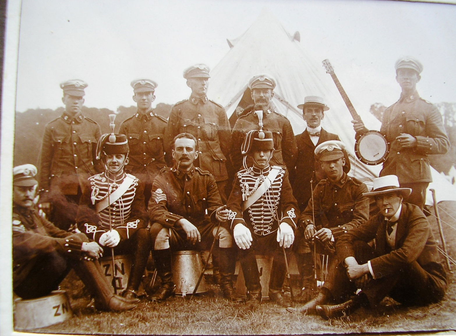 Fatigue party of New Zealanders of ‘D’ Squadron (British African) King’s Colonial’s with visitors from ‘D’ (Chesham) Squadron, The Buckinghamshire Imperial Yeomanry (Royal Buckinghamshire Hussars) and civilians in 1903. The 'D NZ' painted on the upturned pales designate these New Zealand Troopers as belonging to ‘D’ Squadron.  ‘D’ Squadron was the British African Squadron but the 3rd Troop was comprised of the New Zealanders who had originally been part of ‘C’ Squadron (Australasian) in 1902. The Troopers (front left and front third from left) are still wearing the ‘C’ Squadron (Australasian) headdress badge (and an NCO’s arm badge, Figure 151) prior to the fern badge of the New Zealand Squadron being introduced in 1904. Other Troopers (rear left-hand two and front right) are wearing the ‘D’ Squadron (British African) headdress badge. There is a very good description of the Full Dress uniform of the Royal Bucks Hussars (not Imperial) in 'The Yeomanry Force at the 1911 Coronation' by R. J. Smith and R. G. Harris (Picton Publishing, Chippenham, 1988) which matches the uniform worn by the two Buckinghamshire Imperial Yeomanry. A regulation or low pattern Hussars busby with scarlet bag, white lines and white plume. Jacket invisible green with scarlet collar and white chest braiding. Dark blue overalls with double scarlet stripes and white wrist gloves.