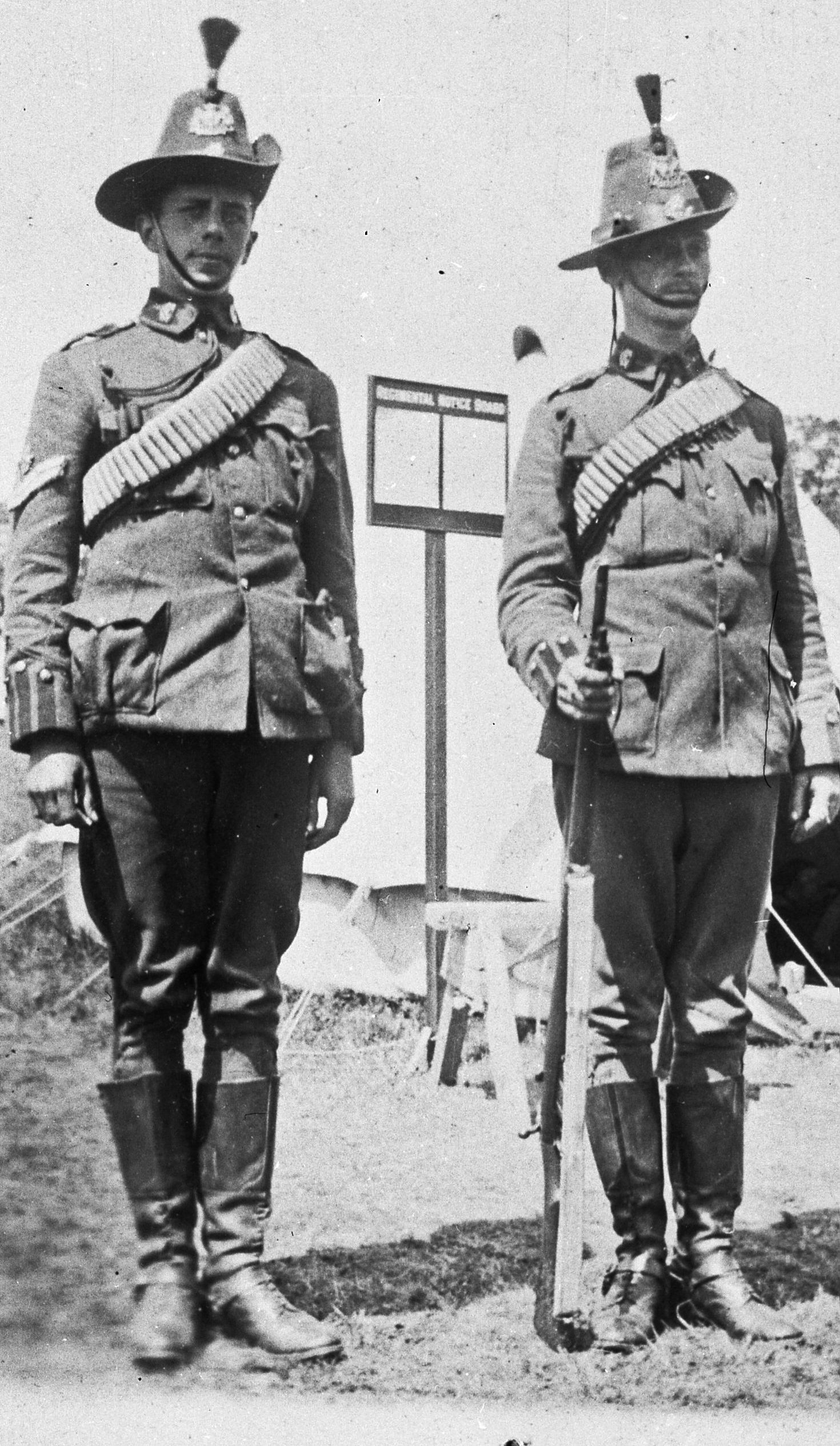 a Corporal and a Private of ‘B’ Squadron (British American) of the 4th County of London (King’s Colonials) Imperial Yeomanry in Full Dress uniform mounting guard whilst on annual summer camp in Sidcup, London in 1902. They are wearing cloth leggings and are equipped with a Boer War pattern, 50-round Mills equipment webbing bandolier and a .303 Small Magazine Lee Enfield Mark 1 rifle.