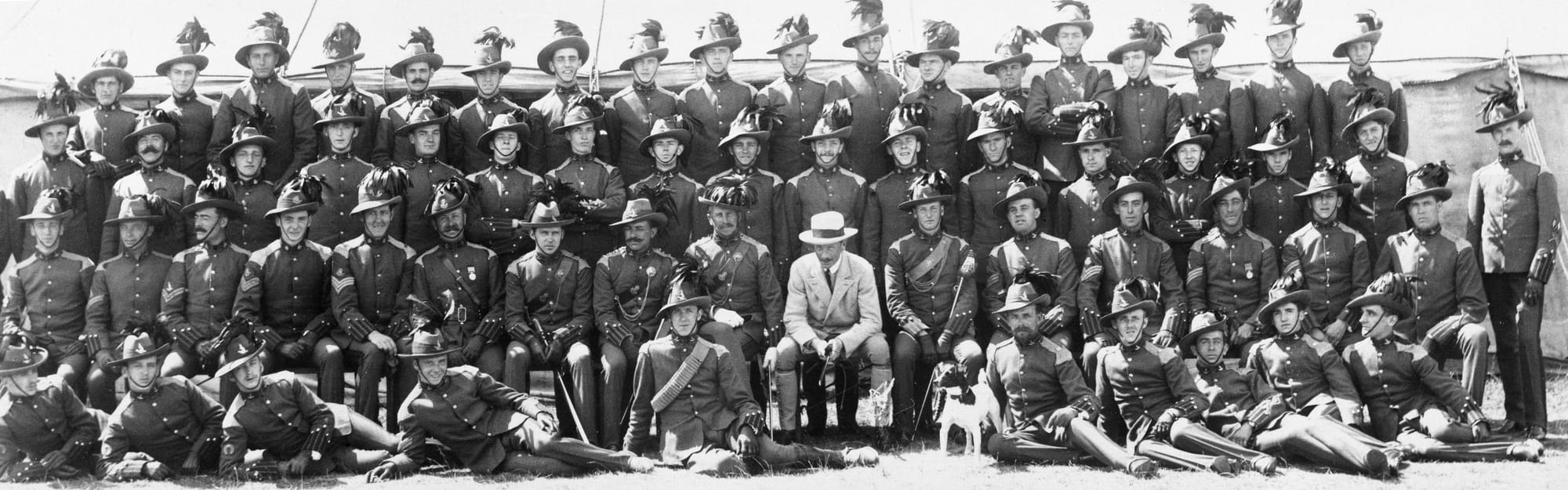 Group photograph of members of 'C' Squadron (Australian) and 'B' Squadron (British American) of the King's Colonials wearing Full Dress uniform with a variety of personalised styles of the 'Sombrero' hat circa 1907 (Courtesy David Knight). The Kangaroo and Beaver headdress and matching collar badges of 'C' and 'B' Squadrons, respectively are shown being worn. The NCOs are also wearing the Kangaroo and Beaver headdress badges as an arm badge above their chevrons. The Staff Officer is wearing the second pattern Regimental badge with matching collars.