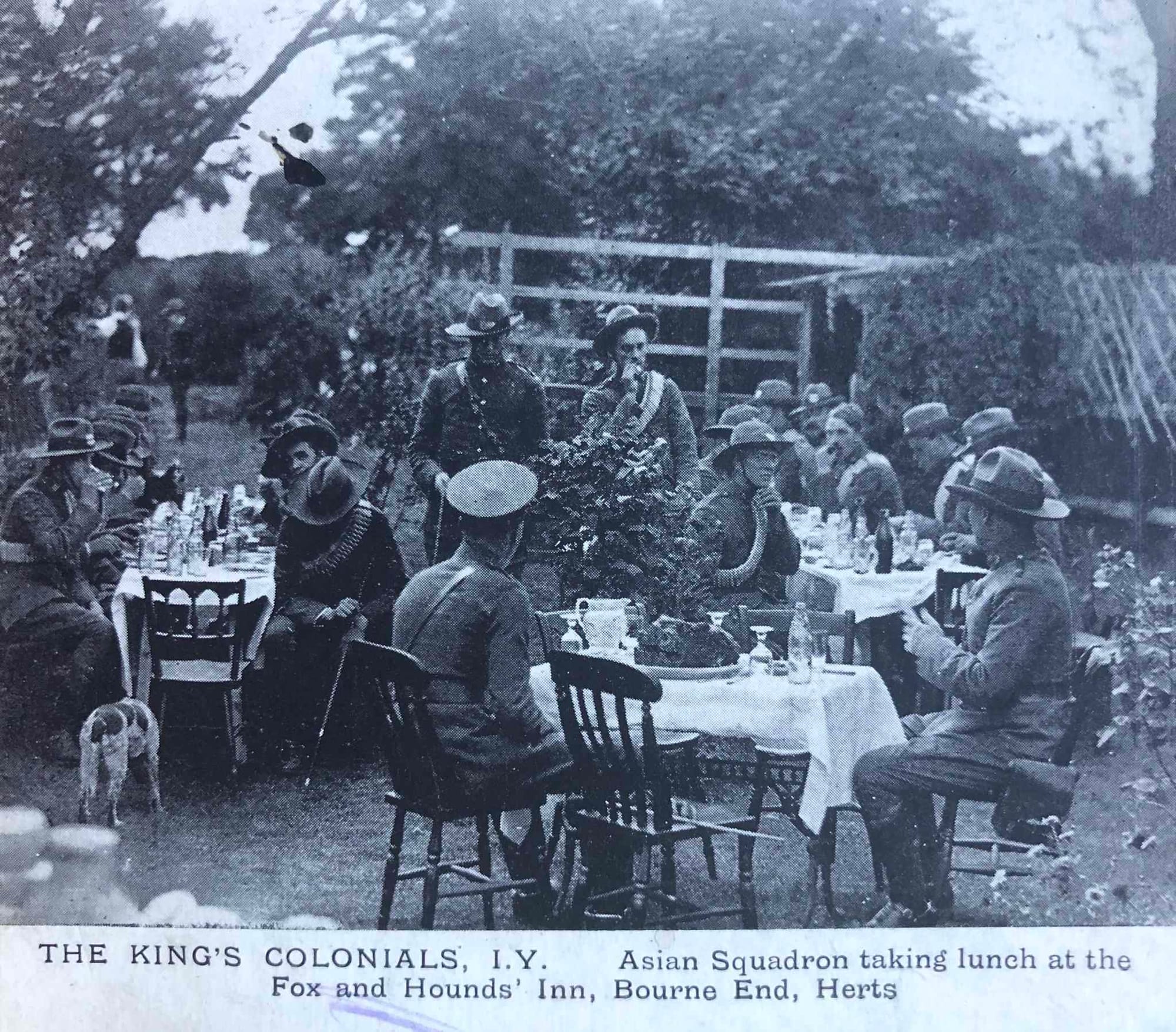 Postcard taken by J. T. Newman, a Berkhamsted photographer depicting members of 'A' (British Asian) Squadron of the King's Colonials at lunch at the Fox and Hounds Inn, Bourne End, Hertfordshire, posted in 1908. At the time the Inn was owned by a Corporal B. Ball of the King's Colonials. (The Fox and Hounds Inn was first licensed in 1863 and was demolished some time after 1940. The Bourne End area is now designated as being in Bedfordshire). They are wearing Service Dress uniforms and the thin (scarlet) cord straps are clearly visible. The Officer (with pouch belt) standing on the left is wearing a New Zealand headdress badge whereas the others are all wearing British Asian headdress badges and matching collar badges. The seated figure to the immediate left of this Officer is wearing an 'A' (British Asian) Squadron headdress badge as an NCO's arm badge. Colonel Lionel James is seated with his back to the camera and is wearing a field service cap. The Other Ranks are all Service Dress tunics with bandoliers and Full Dress Sombrero pattern headdress without plumes.