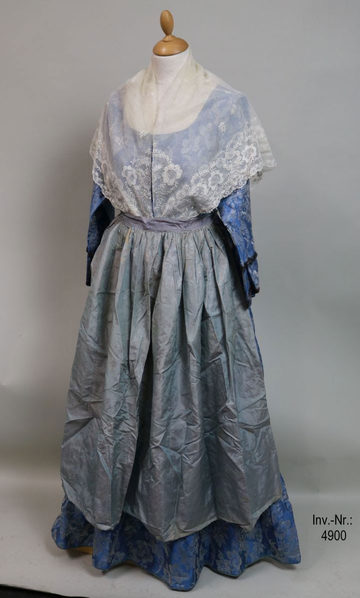 Tracht aus Zuckmantel, weiße Spitzenhaube mit rosa Bändern, hellblaues Kleid mit Webmuster, taubenblaue Schürze, weißes Sptizenschultertuch mit Stickerei.