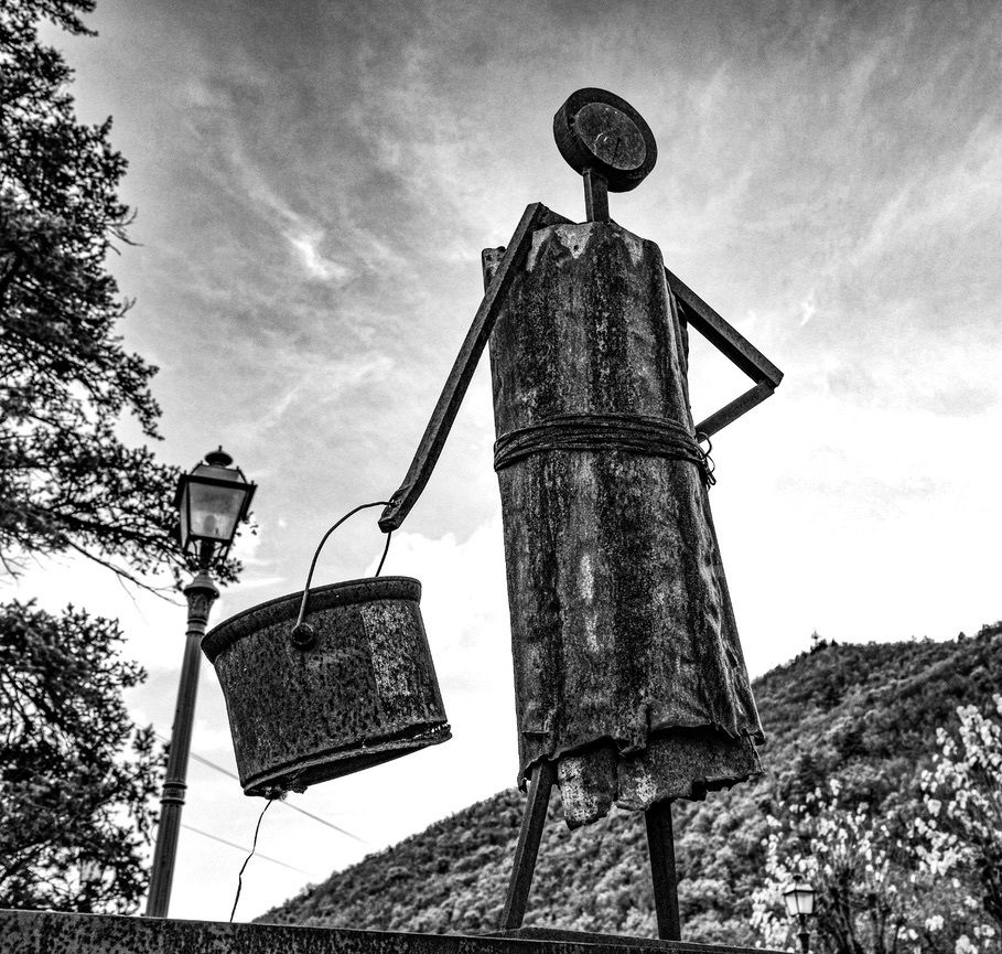 Donna con Secchio  di Roberto Coccoloni. “Donna con secchio”, "Bimbo felice" (Roberto Coccoloni) Le opere di Roberto Coccoloni sono sculture oneste, pulite, emotive. Chi si concede il tempo di stare con loro, si sente invaso di una certa dolcezza malinconica come quando un ricordo prezioso si affaccia e sfuma poi via nel presente affannato. Le forme geometriche, semplici ed essenziali, non rinunciano all’espressività indotta che si aggancia al trascorso quotidiano di ciascuno come un effetto Madeleine. Riportano ad una vita semplice, in un tempo primitivo ma sganciato da riferimenti cronologici, come un passato dell’anima, del soggetto osservante.  Si integrano perfettamente con il paesaggio a cui danno bellezza e valore tanta quanta ne prendono, completandosi con gli elementi intorno.